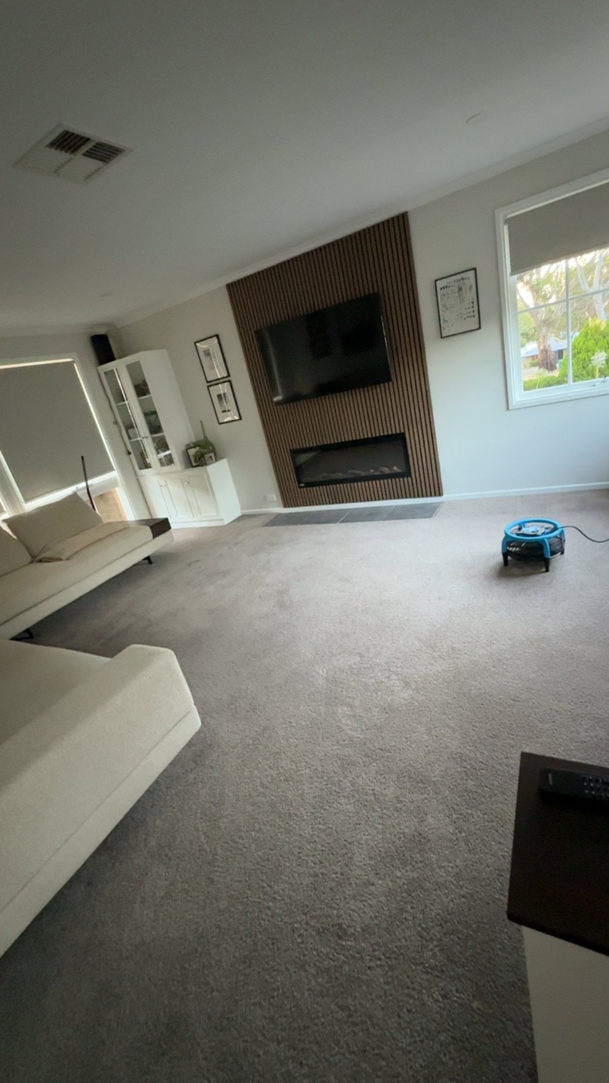 Modern living room with wall-mounted TV, fireplace, white furnishings, and robotic vacuum on gray carpet