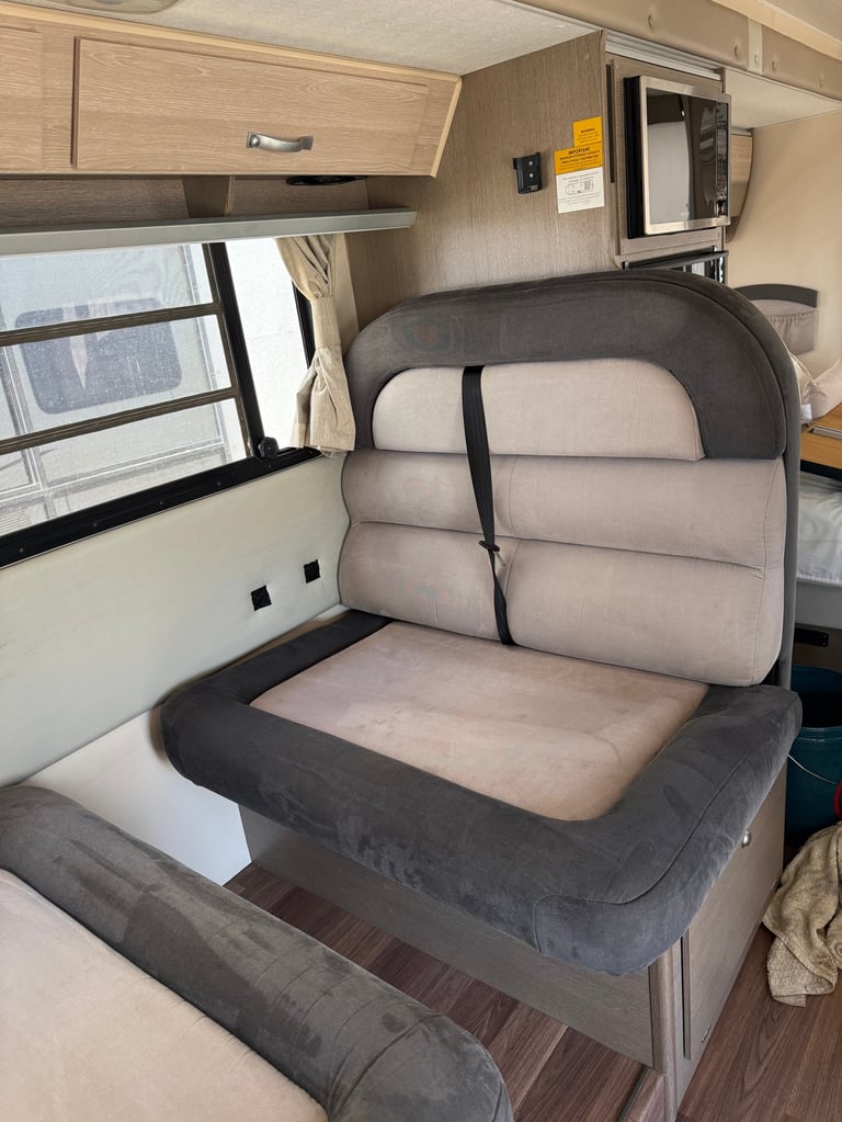 RV interior with convertible sofa bed in gray and beige, window with blinds, microwave above, and wood cabinetry