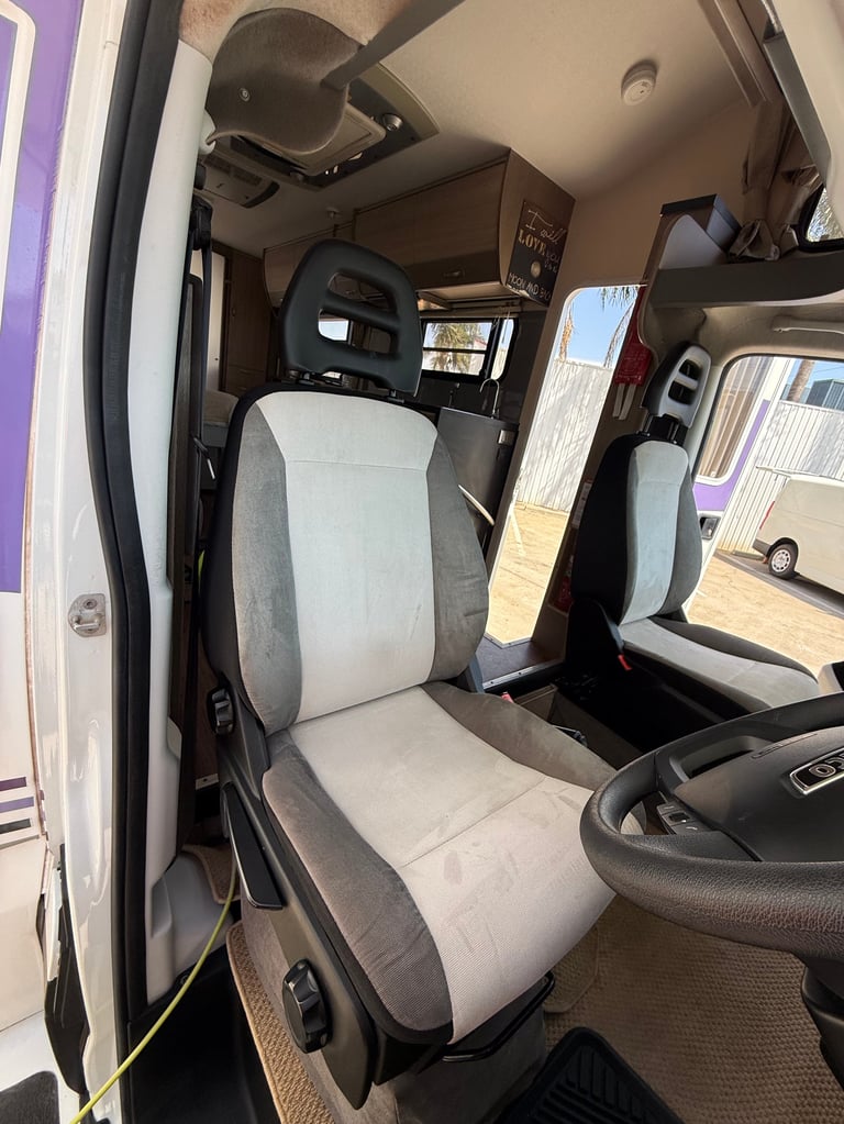 Interior of a commercial truck cabin showing a white and gray drivers seat, dashboard, steering wheel, and windshield view of a sunny outdoor area