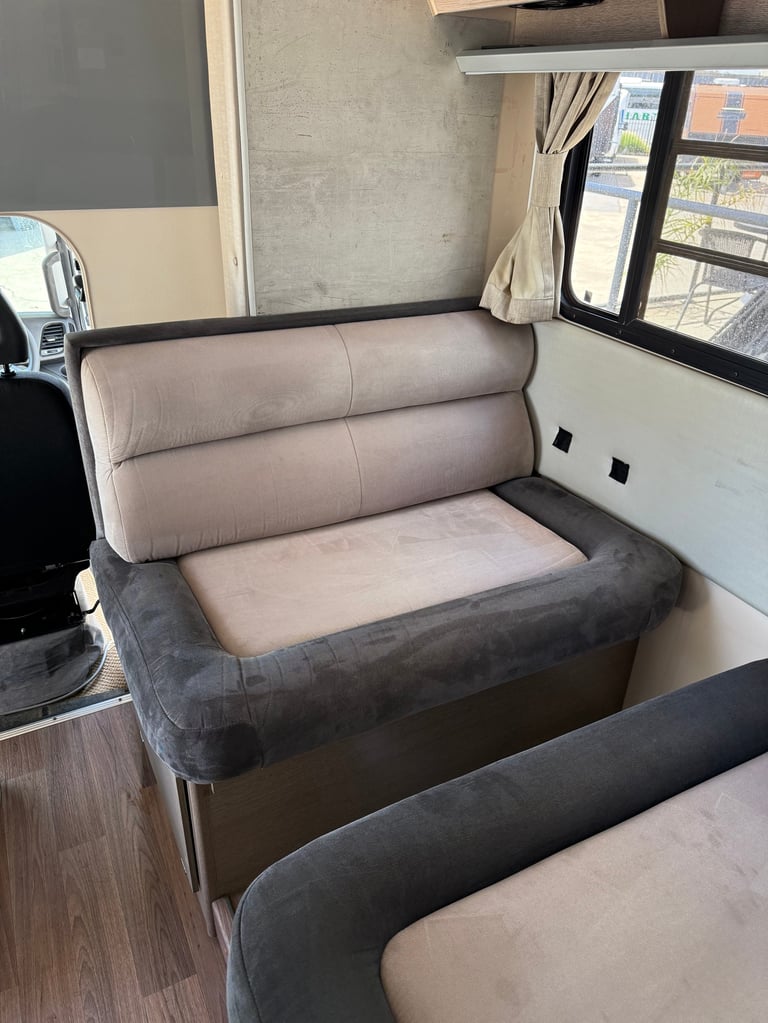 RV interior with beige and gray upholstered seating, window with curtains, and kitchen area in background